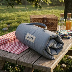 Könnte beinhalten: Eine aufgerollte, gesteppte blaue Picknickdecke mit Tragegriff und einem rechteckigen Aufnäher, der den Namen AMY in dunkelblauer Schrift zeigt. Daneben liegen eine rot-weiß karierte Decke, ein Weidenkorb, zwei Glasflaschen und gefaltete Servietten.