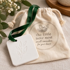 May include: A square white ceramic tile with an embossed leaf design, attached by a dark green satin ribbon to a beige cotton drawstring pouch. The pouch features a printed logo of a bird in a nest and the text, the little note nest.