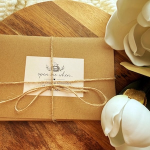 May include: A stack of rectangular brown kraft paper envelopes tied with thin natural jute twine. A white rectangular label is centered on the front, featuring a small bird nest illustration and the text: open me when... you need a reminder of my love.
