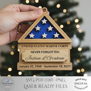 May include: Wooden memorial ornament featuring a flag design with blue and white accents. The ornament includes the text "UNITED STATES MARINE CORPS," "NEVER FORGOTTEN," a name, and dates. The ornament is held by a hand.