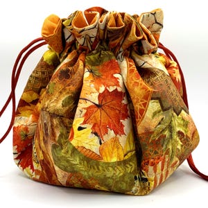 May include: A drawstring bag with a fall-themed print featuring orange, yellow, and green leaves. The bag is made of fabric and has a red drawstring.