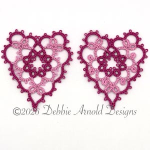 May include: Two heart-shaped lace motifs created with tatting technique in shades of dark magenta and light pink thread. The intricate design features a central circular pattern surrounded by delicate loops and picots. ©2026 Debbie Arnold Designs.