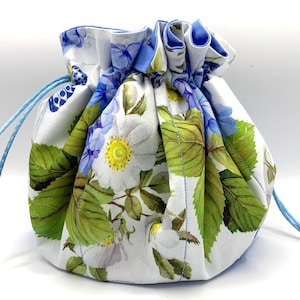 Könnte beinhalten: Ein Stoffbeutel mit Kordelzug auf weißem Grund, bedruckt mit einem botanischen Muster aus grünen Blättern, weißen Blüten mit gelber Mitte und blauen floralen Akzenten. Der Beutel hat einen gerafften Verschluss mit hellblauen Kordeln.