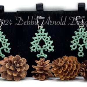 May include: Mint green, handmade, tree-shaped earrings with silver hooks are displayed on a black background. The earrings are made of a delicate, lacy material. Several pine cones are arranged in front of the earrings.
