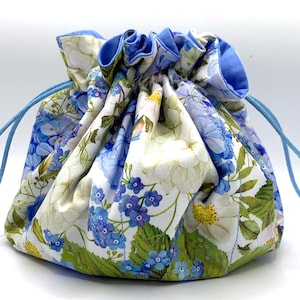 Könnte beinhalten: Ein Stoffbeutel mit Kordelzug und einem Blumenmuster mit blauen, weißen und grünen botanischen Illustrationen. Der Beutel hat einen gerafften Verschluss mit hellblauen Kordeln und ein einfarbig hellblaues Innenfutter aus Stoff.