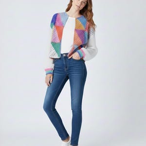May include: A woman wearing a white top, blue jeans, and white sneakers. She is wearing a colorful, geometric-patterned jacket with white sleeves and rainbow-striped cuffs. The jacket features a variety of colors including blue, orange, pink, and green.