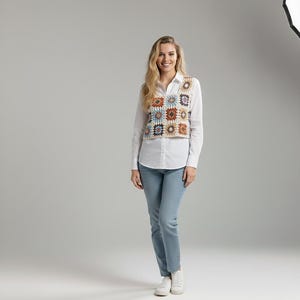 May include: A woman wearing a white button-down shirt, light blue jeans, and white sneakers. She is also wearing a colorful crochet vest with square patterns in shades of brown, blue, and beige.