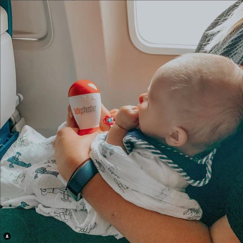May include: A baby being held on an airplane, with a red and white "babyshusher" device. The baby is wearing a bib and wrapped in a white blanket with a blue airplane pattern. A blue fitness tracker is visible on the person's wrist.