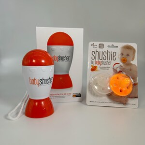 K&ouml;nnte beinhalten: Ein orangefarbener und wei&szlig;er Baby-Shusher mit einem wei&szlig;en Band. Der Shusher ist ein kleines, handliches Ger&auml;t, das ein Shushing-Ger&auml;usch erzeugt, um Babys zu beruhigen. Die Verpackung enth&auml;lt einen wei&szlig;en und orangefarbenen Schnuller mit dem Schriftzug "shushie by babyshusher" auf der Vorderseite.