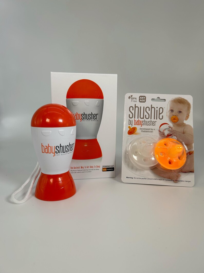 K&ouml;nnte beinhalten: Ein orangefarbener und wei&szlig;er Baby-Shusher mit einem wei&szlig;en Band. Der Shusher ist ein kleines, handliches Ger&auml;t, das ein Shushing-Ger&auml;usch erzeugt, um Babys zu beruhigen. Die Verpackung enth&auml;lt einen wei&szlig;en und orangefarbenen Schnuller mit dem Schriftzug "shushie by babyshusher" auf der Vorderseite.