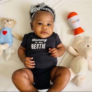 May include: A baby wearing a black onesie with the text "Mommy is my BESTIE" and a leopard print headband. A stuffed bear with a red heart and a stuffed elephant are on either side of the baby. A white and orange baby rattle is also in the image.