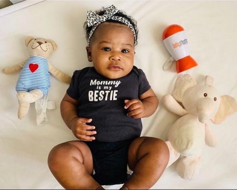 May include: A baby wearing a black onesie with the text "Mommy is my BESTIE" and a leopard print headband. A stuffed bear with a red heart and a stuffed elephant are on either side of the baby. A white and orange baby rattle is also in the image.