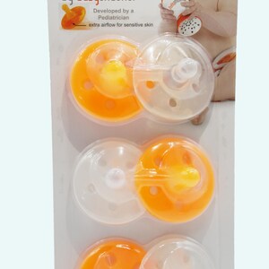 Op de afbeelding: Een verpakking met drie oranje en doorzichtige fopspeen genaamd "shushie by babyshusher". De fopspenen zijn ontworpen met extra luchtstroom voor gevoelige huid. De verpakking bevat een waarschuwing: "Bind de fopspeen niet om de nek van het kind, omdat dit een verstikkingsgevaar oplevert."