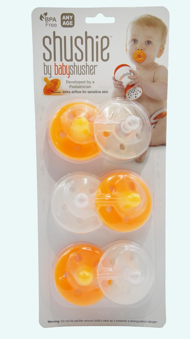 Op de afbeelding: Een verpakking met drie oranje en doorzichtige fopspeen genaamd "shushie by babyshusher". De fopspenen zijn ontworpen met extra luchtstroom voor gevoelige huid. De verpakking bevat een waarschuwing: "Bind de fopspeen niet om de nek van het kind, omdat dit een verstikkingsgevaar oplevert."
