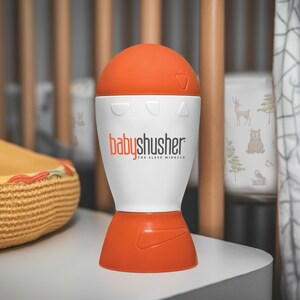 Op de afbeelding: Een witte en oranje baby-shusher geluidsmachine in de vorm van een raket. Het apparaat heeft een witte bovenkant met een rode knop en twee kleinere knoppen. De tekst "babyshusher" en "THE SLEEP MIRACLE" staat op de voorkant van het apparaat.
