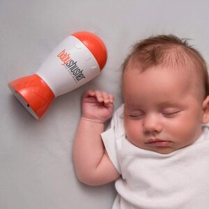 May include: A sleeping baby in a white onesie next to a white and orange baby shusher device. The device has the text "babyshusher" printed on it. The baby is lying on a gray surface.