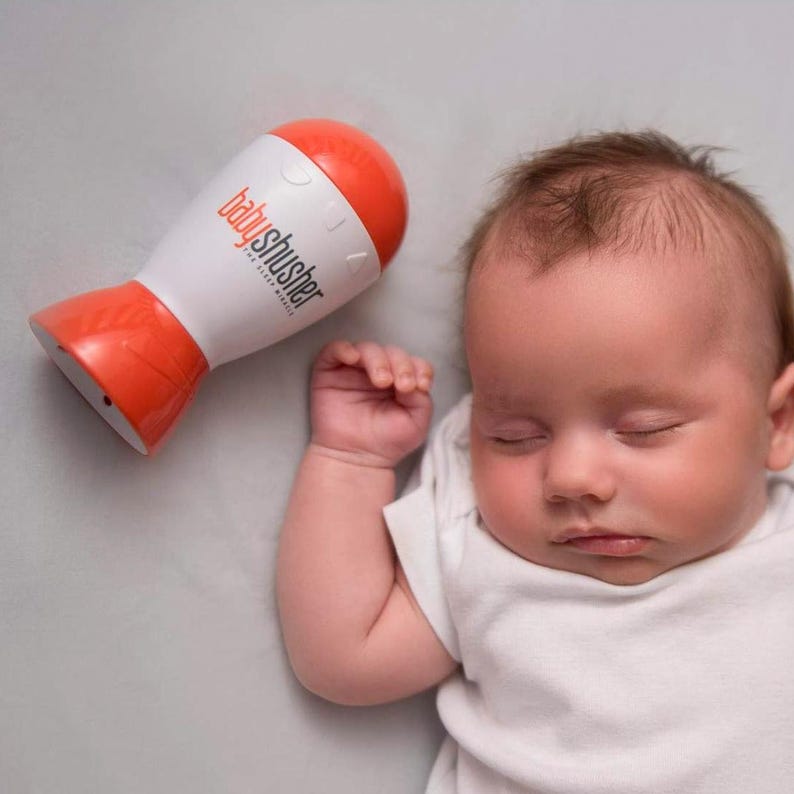May include: A sleeping baby in a white onesie next to a white and orange baby shusher device. The device has the text "babyshusher" printed on it. The baby is lying on a gray surface.
