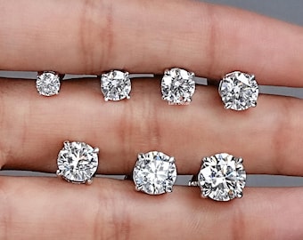 May include: A collection of seven diamond stud earrings of varying sizes, displayed on a hand against a gray background. The earrings are round-cut, with a brilliant sparkle. The sizes range from small to large, showcasing the different carat weights.
