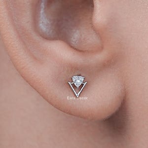 May include: A silver-toned stud earring featuring a triangular frame with a small, clear, faceted stone set at the top point. The earring is positioned in the earlobe and displays the text Ears Decor printed in small white letters below it.