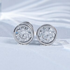 May include: A pair of silver-toned stud earrings featuring round, clear, faceted stones set in a smooth bezel setting. Each earring displays the text Ears Decor in small, white lettering at the base of the metal rim, showcasing a polished finish.
