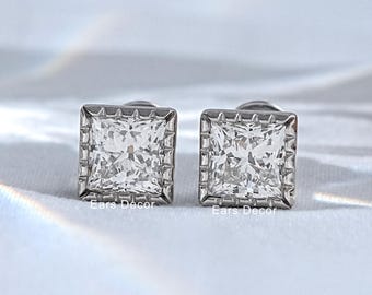 May include: A pair of silver-toned stud earrings featuring square-cut clear stones set in a metallic frame with a textured border. The earrings are positioned side-by-side, displaying a faceted surface that reflects light, with the text Ears Décor visible.