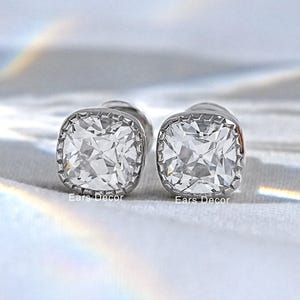 May include: A pair of silver-toned stud earrings featuring square-cut clear stones with faceted surfaces. Each earring is set in a metallic frame with a subtle notched edge detail. The text Ears Decor is printed in small white letters below each stone.