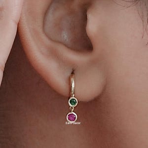 Könnte beinhalten: Goldene Creolen mit einem baumelnden Anhänger aus grünen und roten Edelsteinen. Die Ohrringe sind an einem Ohr einer Person zu sehen.