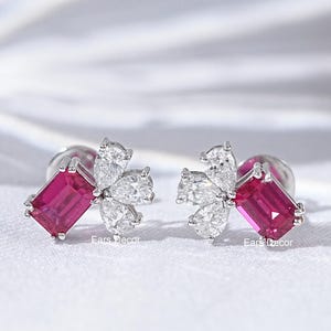 Peut inclure: Une paire de boucles d'oreilles à clous avec une pierre précieuse rose foncé rectangulaire et un groupe de pierres claires en forme de fleur. Les boucles d'oreilles sont posées sur une surface blanche.