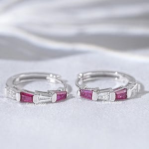 Puede incluir: Un par de pendientes de aro de plata con una fila de pequeñas piedras preciosas rojas y transparentes.