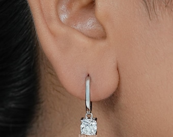 May include: A silver-toned hoop earring featuring a single, clear, cushion-cut gemstone drop. The earring is secured through the earlobe with a hinged closure, showcasing a polished metallic finish and a faceted stone that reflects light.