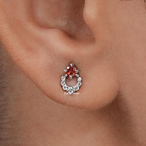 May include: A pair of silver stud earrings with a red gemstone in the centre and a circle of smaller white gemstones around it.