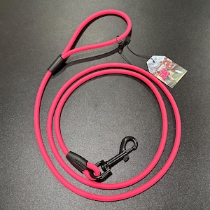 May include: A vibrant pink dog lead with a black metal clasp and handle loop. The lead is made of a flexible, durable material and includes a small tag with an image. Designed for pet owners.