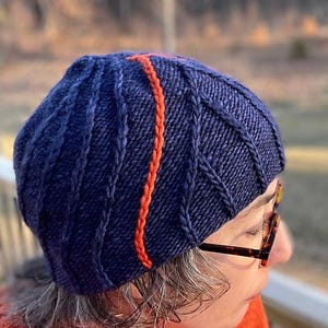 May include: A navy blue knitted hat with a thick orange stripe down the side. The hat has a cable knit pattern.