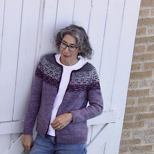 May include: A woman wearing a purple and grey patterned cardigan with a white t-shirt underneath. She is standing in front of a white wooden door.