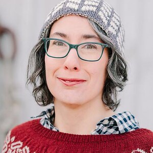 May include: A woman wearing a gray and white patterned knit hat, green glasses, and a red sweater with a white geometric pattern.
