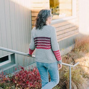 May include: A gray sweater with red and pink horizontal stripes across the chest and on the cuffs. The person is wearing blue jeans and black boots. The person is walking down a set of concrete stairs.