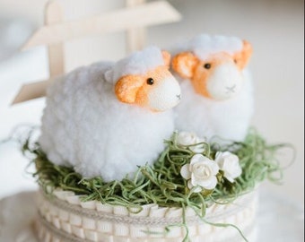 Può includere: Decorazione per torta con due pecore bianche con dettagli arancioni, su erba sintetica verde e fiori bianchi. La base è color crema con dettagli a nastro e una piccola staccionata in legno.