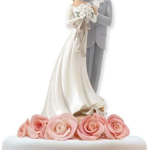 K&ouml;nnte beinhalten: Ein wei&szlig;er Hochzeitstortenaufsatz mit einer Braut- und Br&auml;utigamfigur. Die Braut tr&auml;gt ein wei&szlig;es Kleid und einen Schleier, der Br&auml;utigam einen grauen Anzug. Das Paar steht auf einem Sockel, der mit rosa Rosen verziert ist.