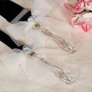 May include: A set of two white crystal cake servers with blue flower accents and white tulle bows. The servers are on a white background.