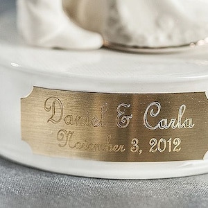 May include: A white ceramic wedding cake topper base with a gold plaque. The plaque is engraved with the names "Daniel & Carla" and the date "November 3, 2012".
