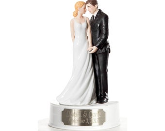 May include: A white porcelain wedding cake topper featuring a bride and groom in a romantic pose. The bride is wearing a white gown and the groom is wearing a black suit. The base of the topper is white with a silver plaque that reads "David & Carla November 3, 2012".