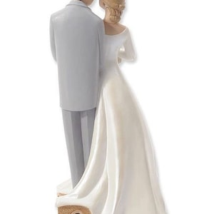 K&ouml;nnte beinhalten: Ein wei&szlig;er Hochzeitstortenaufsatz mit einer Braut und einem Br&auml;utigam in traditioneller Hochzeitspose. Die Braut tr&auml;gt ein wei&szlig;es Kleid mit einer Blumenkrone und der Br&auml;utigam einen grauen Anzug. Das Paar steht auf einem kleinen, kirchenf&ouml;rmigen Sockel.