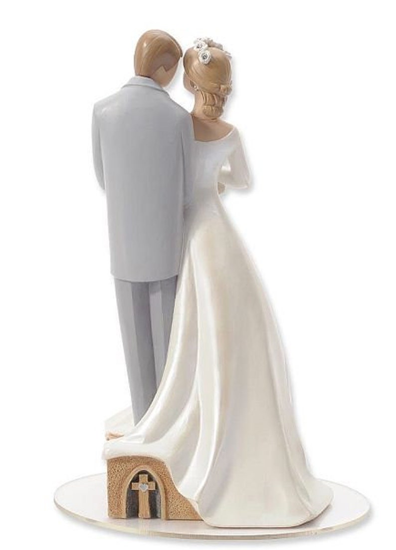 K&ouml;nnte beinhalten: Ein wei&szlig;er Hochzeitstortenaufsatz mit einer Braut und einem Br&auml;utigam in traditioneller Hochzeitspose. Die Braut tr&auml;gt ein wei&szlig;es Kleid mit einer Blumenkrone und der Br&auml;utigam einen grauen Anzug. Das Paar steht auf einem kleinen, kirchenf&ouml;rmigen Sockel.