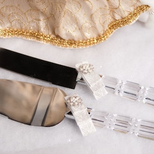 May include: A silver cake knife and server set. The knife has a serrated blade, and the server has a wide, flat blade. Both have clear, faceted handles and are adorned with white lace and floral embellishments. A gold and cream fabric is in the background.