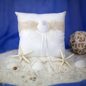 May include: A white ring pillow with a seashell and ribbon embellishment. The pillow is decorated with a beige sequined band and a white bow. Two starfish with rhinestone accents and seashells are on a bed of sand.