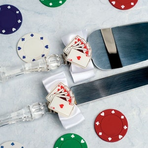 May include: A pair of crystal cake servers with white satin bows and playing card accents. The servers are decorated with a stack of playing cards with red hearts. The servers are surrounded by poker chips in various colors.