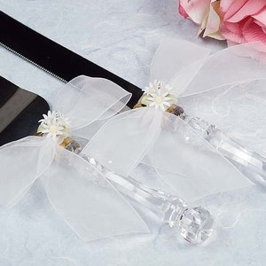 May include: A wedding cake knife and server set. The blades are black, and the handles are clear with a faceted design. White sheer bows and small white flower accents adorn the handles, creating an elegant look.
