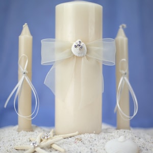 May include: A set of ivory-colored wedding candles. A large pillar candle is adorned with a sheer bow and a seashell embellishment. Two smaller taper candles flank the pillar candle. The scene includes starfish and a seashell on a sandy surface.