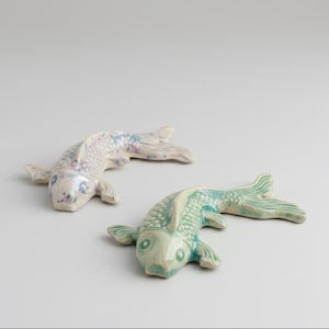 Op de afbeelding: Twee keramische koi-visbeeldjes, één met een bleek lavendelkleurig en wit glazuur met subtiele blauwe accenten, en de andere afgewerkt in een levendig zeegroen glazuur. Beide vissen tonen gedetailleerde schubstructuren en gebeeldhouwde vinnen.