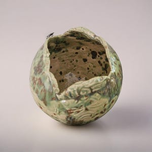 May include: A handcrafted ceramic bowl with a unique, organic shape. The bowl features a mottled green and brown glaze with a textured surface. The rim has an uneven, sculpted edge, adding to its artistic appeal. The interior has dark spots.